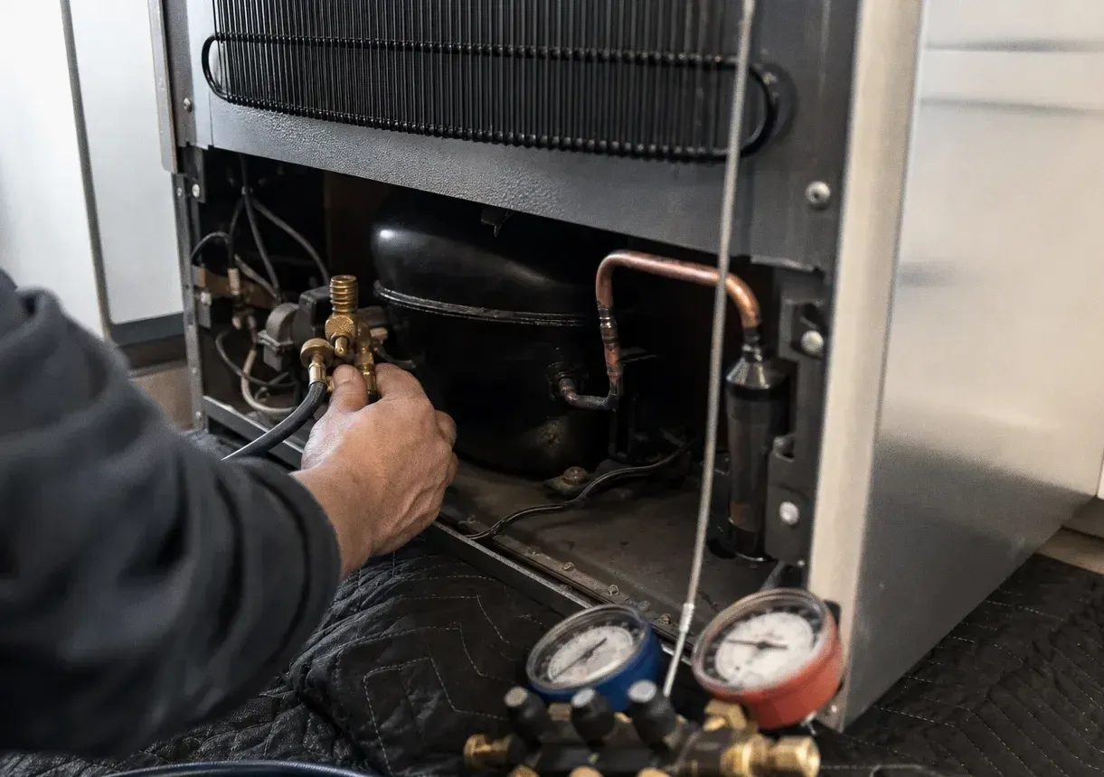 Rear of a refrigeration cabinet opened to its compressor and copper lines, with a twin-dial manifold gauge set in the foreground