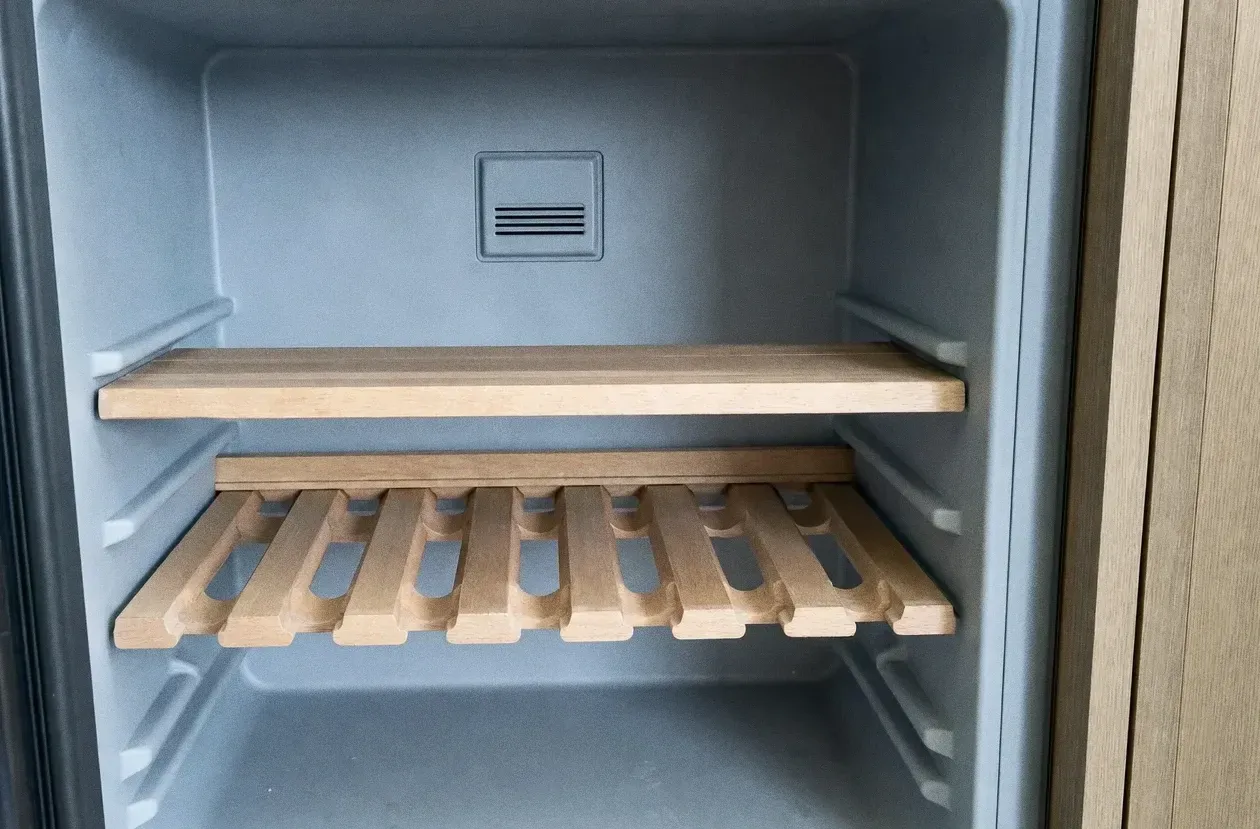 Empty pale refrigerator interior with one glass shelf, one wooden slatted shelf and a return-air grille on the back wall